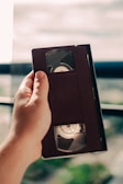 A hand holding a VHS tape in front of a blurred background, possibly near a window with light streaming in.