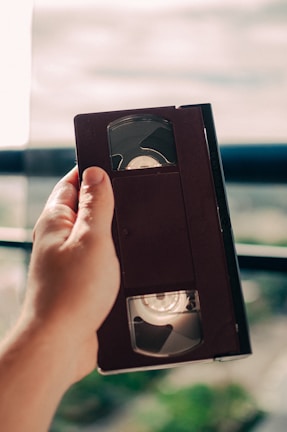 A hand holding a VHS tape in front of a blurred background, possibly near a window with light streaming in.