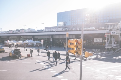 A bustling urban scene near the Barcelona Sants station with pedestrians walking, cars parked, and a row of white tents possibly indicating a market or event. There is a bright, sunny atmosphere with a distant building and terrace full of cars. A traffic light partially obstructs the foreground, adding to the busy city vibe.