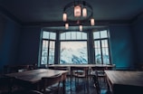 A spacious room with several wooden tables and chairs arranged around the space. Large windows allow a breathtaking view of snow-capped mountains. The room's ambiance is enhanced by stylish ceiling lights that cast a warm glow.