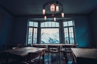 A spacious room with several wooden tables and chairs arranged around the space. Large windows allow a breathtaking view of snow-capped mountains. The room's ambiance is enhanced by stylish ceiling lights that cast a warm glow.