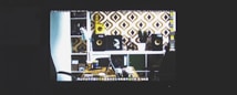 A computer monitor displaying a desktop with various icons is visible. The wallpaper features a modern design with geometric patterns in brown and white. The image includes a desk setup with a laptop, a desk lamp, a potted plant, and speakers.