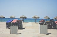 Sun loungers stacked in neat piles on a sandy beach. Several thatched umbrellas line the edge of the sand near the shimmering blue ocean, with a wooden walkway leading to the water.