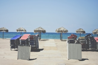 Sun loungers stacked in neat piles on a sandy beach. Several thatched umbrellas line the edge of the sand near the shimmering blue ocean, with a wooden walkway leading to the water.