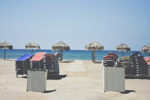 Sun loungers stacked in neat piles on a sandy beach. Several thatched umbrellas line the edge of the sand near the shimmering blue ocean, with a wooden walkway leading to the water.