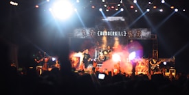 A live concert with a rock band performing on stage. The stage is illuminated with bright lights and smoke effects. The band's name is displayed prominently on a large banner behind the drummer. The audience is visible in the foreground, some holding up their phones or hands.