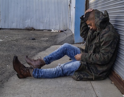 A person wearing a camo jacket and blue jeans sits on the ground against a corrugated metal wall. Their eyes are closed, and they hold their head with one hand, suggesting a moment of contemplation or distress. The environment appears industrial, with a cement path and some debris scattered around.