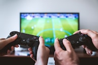 Two hands holding game controllers positioned in front of a television screen displaying a soccer game. The scene suggests a multiplayer gaming session with intense focus on the match.
