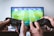 Two hands holding game controllers positioned in front of a television screen displaying a soccer game. The scene suggests a multiplayer gaming session with intense focus on the match.