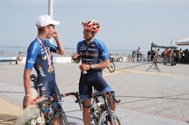 Two cyclists wearing matching blue cycling gear are standing with their bicycles on a paved area near the sea. One is holding a bouquet of flowers and wearing a white cap with medals around his neck, while the other has an orange helmet and is gesturing with a bottle in hand. In the background, there are more people with bicycles and structures suggesting an outdoor gathering or event by the waterfront.