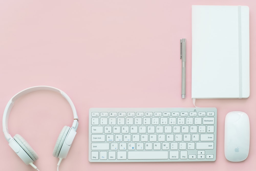 A flatlay with a white notebook, keyboard, headphones and mouse on a pink surface