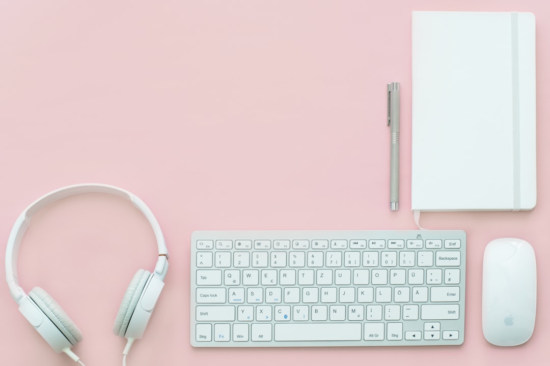 A flatlay with a white notebook, keyboard, headphones and mouse on a pink surface