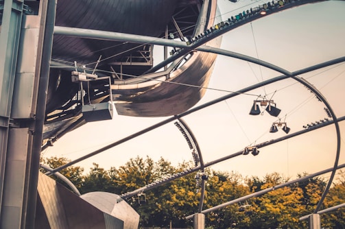 A complex structure composed of curved metallic beams and panels, likely part of a modern architectural design. The foreground consists of metal framework and light fixtures, while the background reveals green trees under a warm, clear sky.