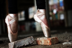 Ballet dancer's feet in pointe shoes balancing on bricks in a dimly lit, possibly industrial setting. The focus is on the dancer&rsquo;s elegant pose and the texture of the bricks.