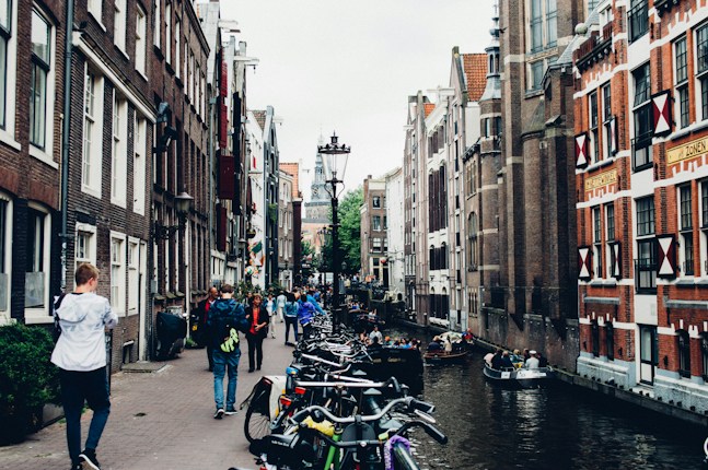 A lively city street along a canal with people walking and biking. Historic buildings with traditional architecture line both sides. Bicycles are parked along the canal's edge, and several boats with people are navigating the water. The sky is overcast, adding a mellow ambiance to the scene.