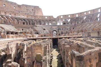 An ancient amphitheater with multiple tiers of stone seating surrounding a central arena. The structure is partially ruined, revealing the underground network of passages. Tourists are visible walking on elevated platforms, observing the historical site.