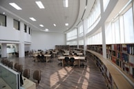 A spacious library with high ceilings, large windows, and numerous bookshelves along the walls. The room is filled with several tables and chairs, arranged neatly, creating a bright and welcoming study environment.