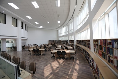 A spacious library with high ceilings, large windows, and numerous bookshelves along the walls. The room is filled with several tables and chairs, arranged neatly, creating a bright and welcoming study environment.