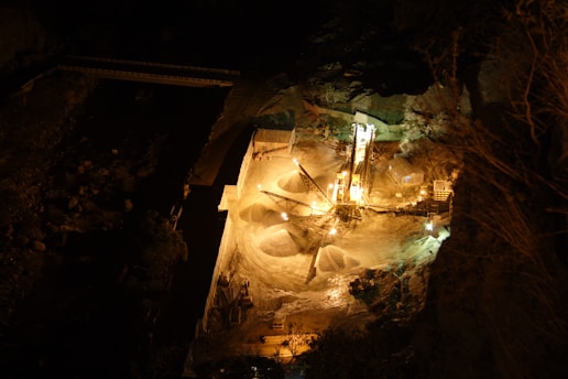 The image depicts a nighttime view of an industrial site, possibly a quarry or a construction area, illuminated by artificial lights. The setup includes machinery and piles of materials, and the surrounding area is dark, enhancing the contrast between the brightly lit equipment and the shadows.