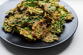 A black plate filled with vibrant green and golden-brown kale chips. The crispy leaves have a textured surface, indicating they are seasoned or baked.
