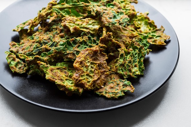 A black plate filled with vibrant green and golden-brown kale chips. The crispy leaves have a textured surface, indicating they are seasoned or baked.