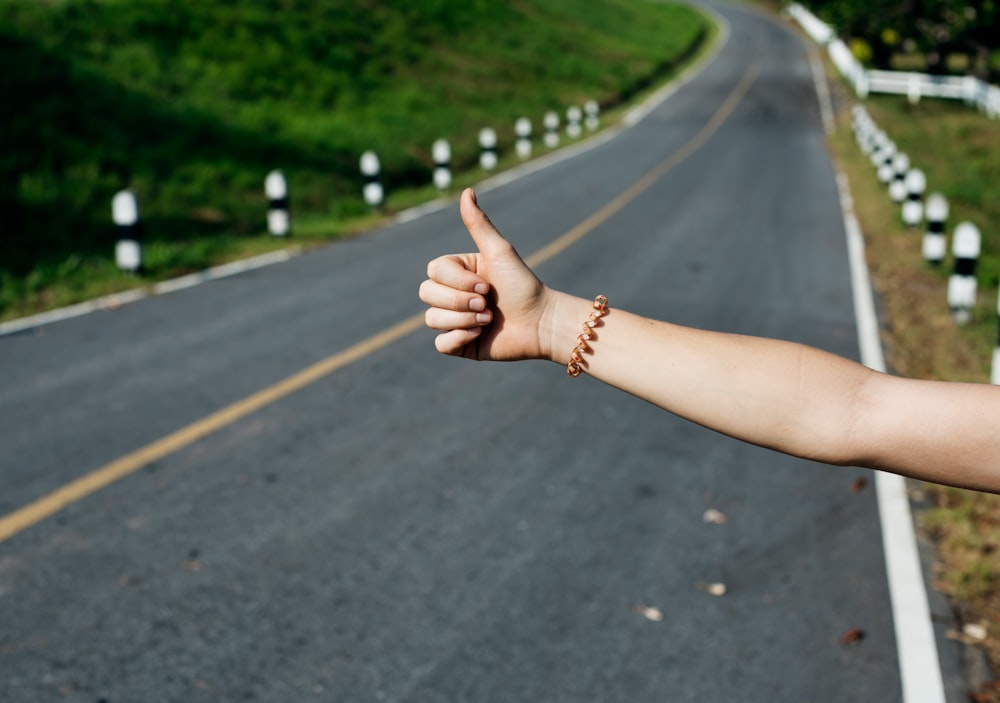 Person's arm with bracelet displaying hitch hiking sign near asphalt road