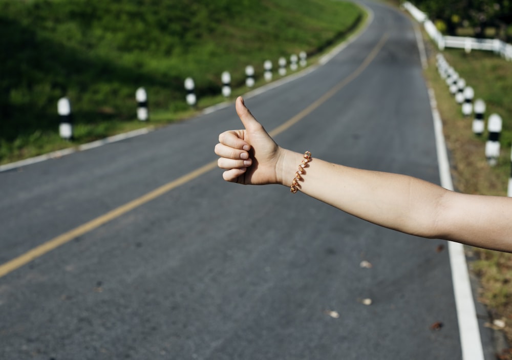 Person's arm with bracelet displaying hitch hiking sign near asphalt road