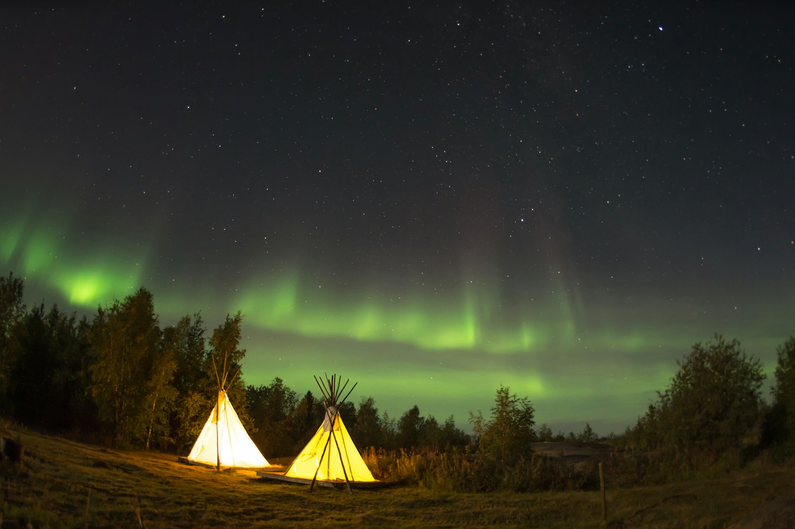 Lighted tipi tent in a snowy forest under green northern lights near Yellowknife