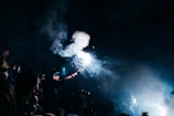 A person is holding a bright flare surrounded by smoke, creating an intense light in a dark, crowded setting. The scene appears energetic and chaotic, with blurred figures and raised hands visible.