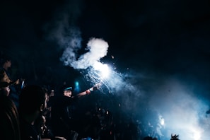 A person is holding a bright flare surrounded by smoke, creating an intense light in a dark, crowded setting. The scene appears energetic and chaotic, with blurred figures and raised hands visible.