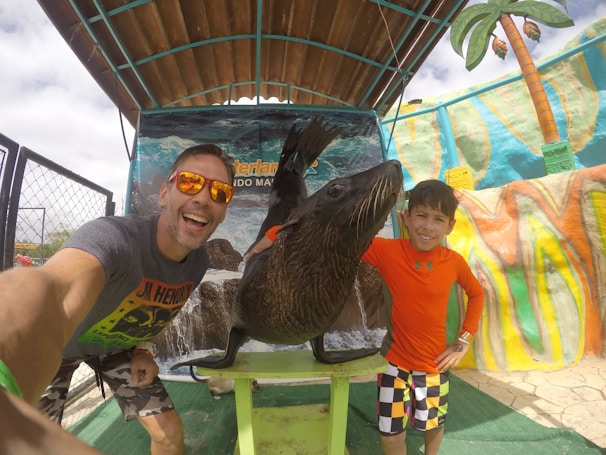 Two people are posing with a sea lion under a shelter at what seems to be a zoo or marine park. The person on the left is wearing sunglasses and a gray t-shirt, while the person on the right is in an orange long-sleeve shirt with colorful shorts. The background includes a vivid painted mural and a fake palm tree.