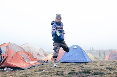 A young person in a blue jacket is jumping energetically among colorful tents on a misty day. The grassy field is sprinkled with several camping tents, primarily in shades of red, blue, and beige. The background is foggy, giving a sense of early morning or late afternoon chill.