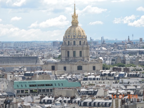 A cityscape with a prominent dome featuring intricate golden details, surrounded by a multitude of traditional buildings with gray and beige tones. The sky is partly cloudy, adding depth to the urban setting.