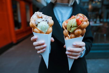 A person is holding two bubble waffles in white paper cones. Each waffle is filled with scoops of ice cream; one has green and white ice cream with chocolate drizzle and the other has green ice cream with strawberries on top. The background appears to be an urban outdoor setting.