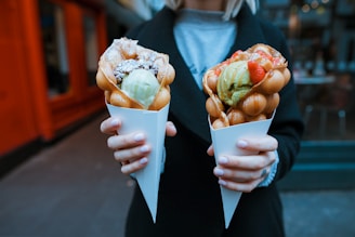 A person is holding two bubble waffles in white paper cones. Each waffle is filled with scoops of ice cream; one has green and white ice cream with chocolate drizzle and the other has green ice cream with strawberries on top. The background appears to be an urban outdoor setting.