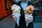 A person is holding two bubble waffles in white paper cones. Each waffle is filled with scoops of ice cream; one has green and white ice cream with chocolate drizzle and the other has green ice cream with strawberries on top. The background appears to be an urban outdoor setting.