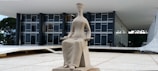 A modern, minimalist sculpture of a seated figure, holding an object, is positioned in an open public space. Behind the sculpture stands a contemporary glass and concrete building with large windows and a flat roof.