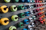 Multiple rows of rolls of colored textile or tape are mounted on spindles attached to a wall. Each roll has a different color, ranging from shades of green, blue, red, yellow, purple, and orange. The rolls are neatly organized and evenly spaced across the wall.