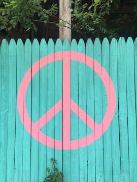 A teal wooden fence features a large pink peace symbol painted on it. There are green leaves and branches hanging over the top edge of the fence, and some foliage is seen growing in front of it.