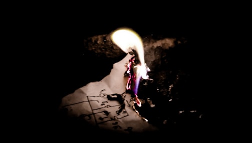 A piece of paper with scribbled text and diagrams is ignited, with vibrant flames and visible embers consuming it. The background is dark, highlighting the paper and fire.