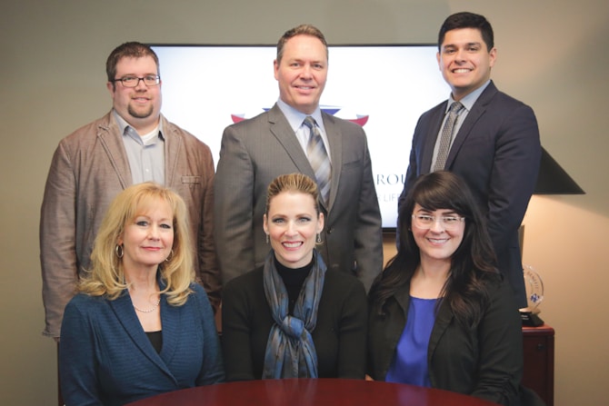 A group of six professionally dressed individuals are posed together in an office setting. Three women and three men are standing and sitting, smiling towards the camera. The background features a flat-screen monitor and a table with a lamp and decorative items.