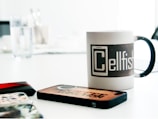 A table is filled with several phone cases and a mug. The mug features a design with the word 'Cellfish' prominently displayed. The table appears to be white, with a glass of water and some utensils visible in the blurred background.