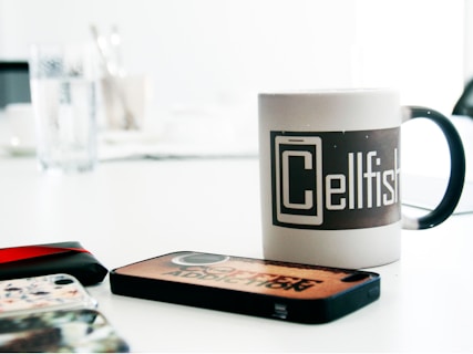 A table is filled with several phone cases and a mug. The mug features a design with the word 'Cellfish' prominently displayed. The table appears to be white, with a glass of water and some utensils visible in the blurred background.