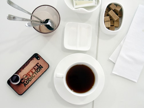 A white table surface displays a setup for enjoying coffee, featuring a cup of dark coffee on a white saucer. To the right, there is a small bowl filled with brown sugar cubes. A clear glass holds three metallic spoons, with an empty dish for condiments nearby. A smartphone with a cover that reads 'Coffee Addiction' lies on the table, and white napkins are placed to the side.