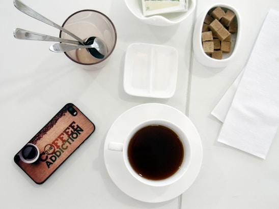 A white table surface displays a setup for enjoying coffee, featuring a cup of dark coffee on a white saucer. To the right, there is a small bowl filled with brown sugar cubes. A clear glass holds three metallic spoons, with an empty dish for condiments nearby. A smartphone with a cover that reads 'Coffee Addiction' lies on the table, and white napkins are placed to the side.