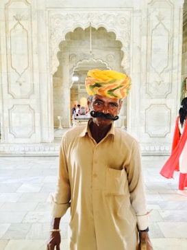 An older man with a large mustache wearing a colorful turban and beige clothing stands in an intricately decorated marble hallway. The background features ornate carvings and arches, lending a historical or cultural atmosphere to the setting.