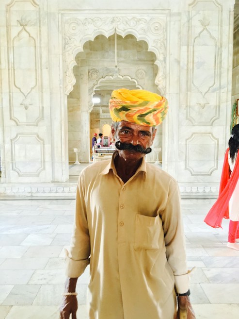 An older man with a large mustache wearing a colorful turban and beige clothing stands in an intricately decorated marble hallway. The background features ornate carvings and arches, lending a historical or cultural atmosphere to the setting.