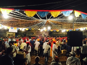 A lively outdoor celebration with a large crowd of people dancing under colorful draped decorations and bright lights. Many individuals are wearing traditional clothing, including colorful turbans, reflecting a festive atmosphere. The setting seems to be a public square or street, with stalls and buildings in the background.