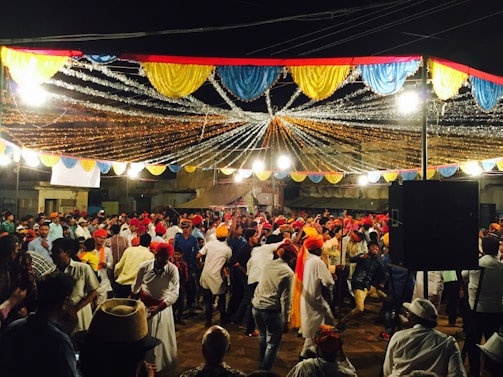 A lively outdoor celebration with a large crowd of people dancing under colorful draped decorations and bright lights. Many individuals are wearing traditional clothing, including colorful turbans, reflecting a festive atmosphere. The setting seems to be a public square or street, with stalls and buildings in the background.
