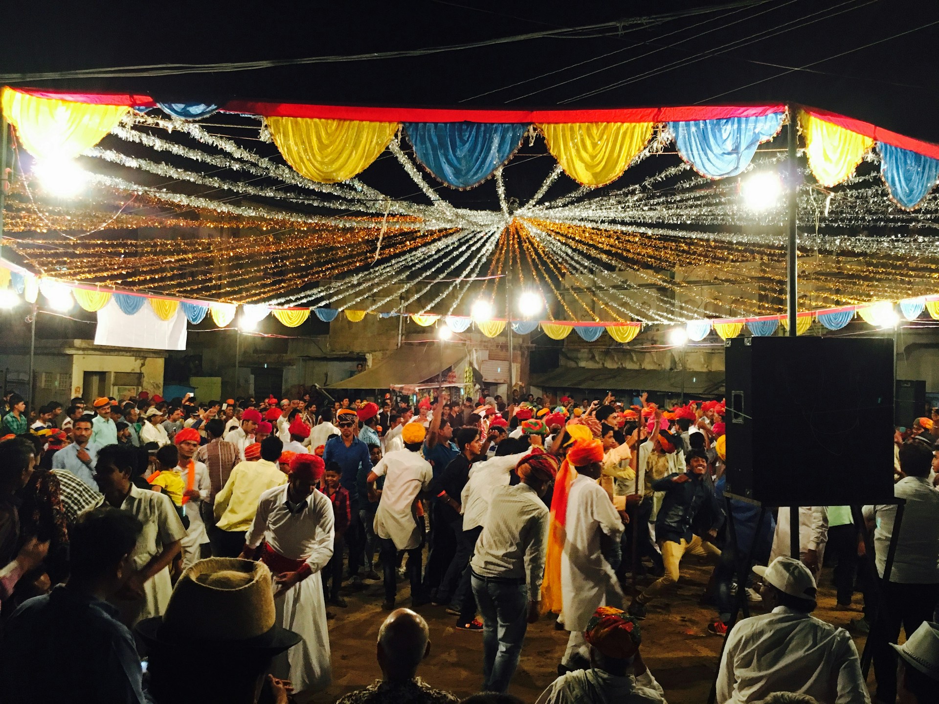 A lively dhol player in traditional Punjabi attire energizing a wedding crowd under colorful umbrella lights.